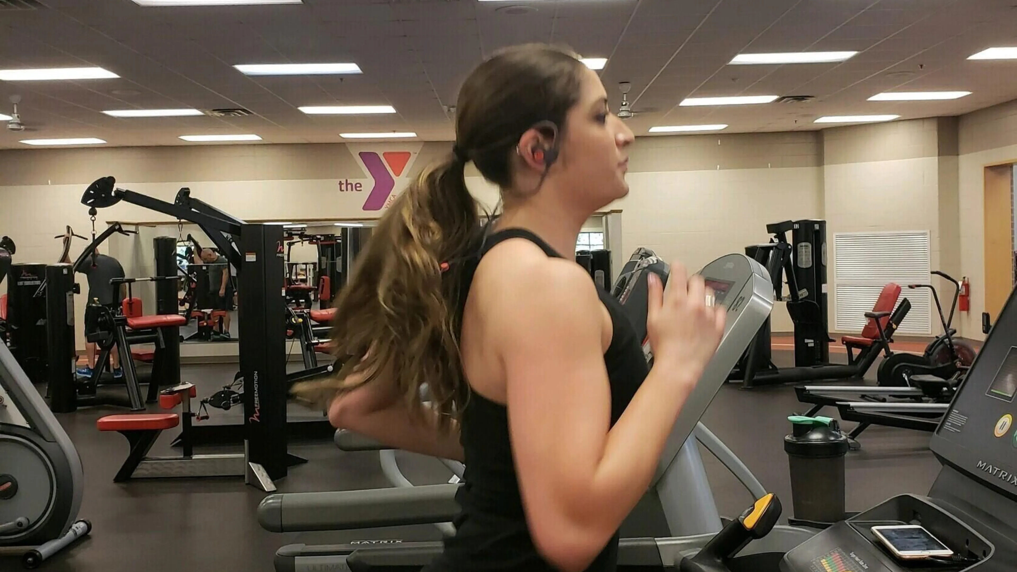 A woman with long brown hair runs on a treadmill in a fitness center; other exercise equipment is visible in the background.