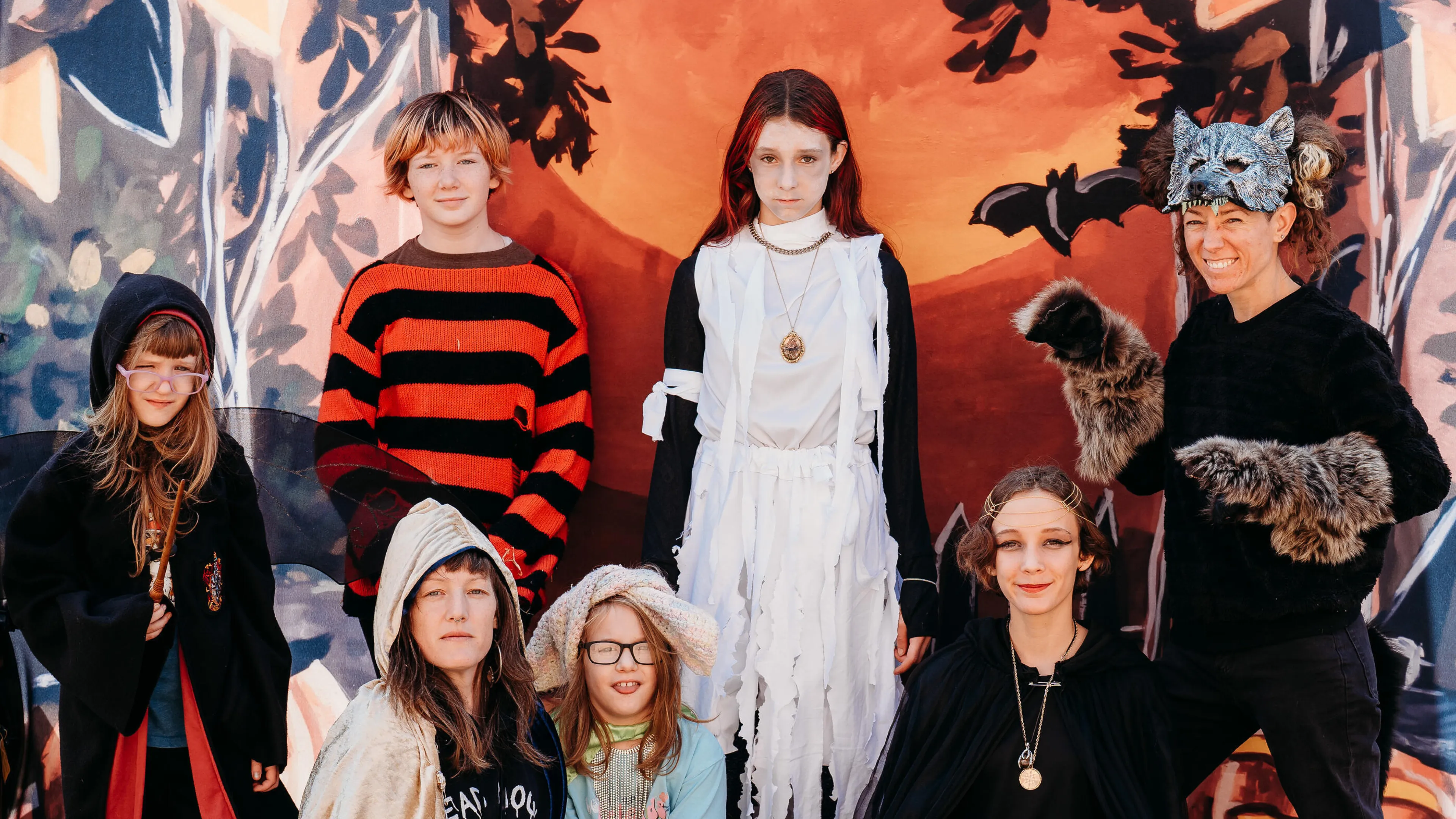 Kids in Halloween costumes pose for a photo; a girl dressed as a ghost, others in fantasy attire,  against a painted backdrop.