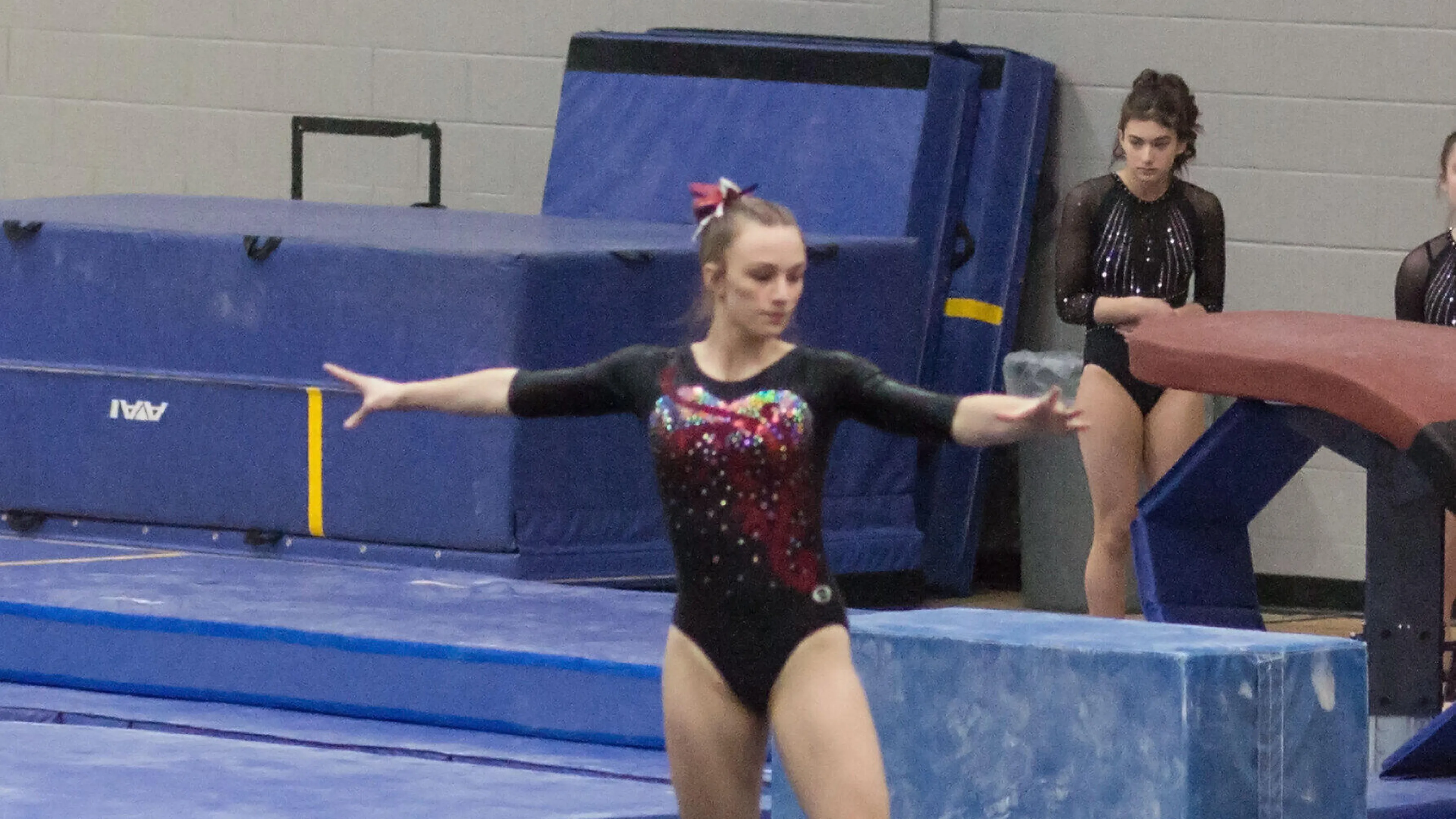 A young gymnast in a black and red leotard performs a floor routine; other gymnasts watch nearby.