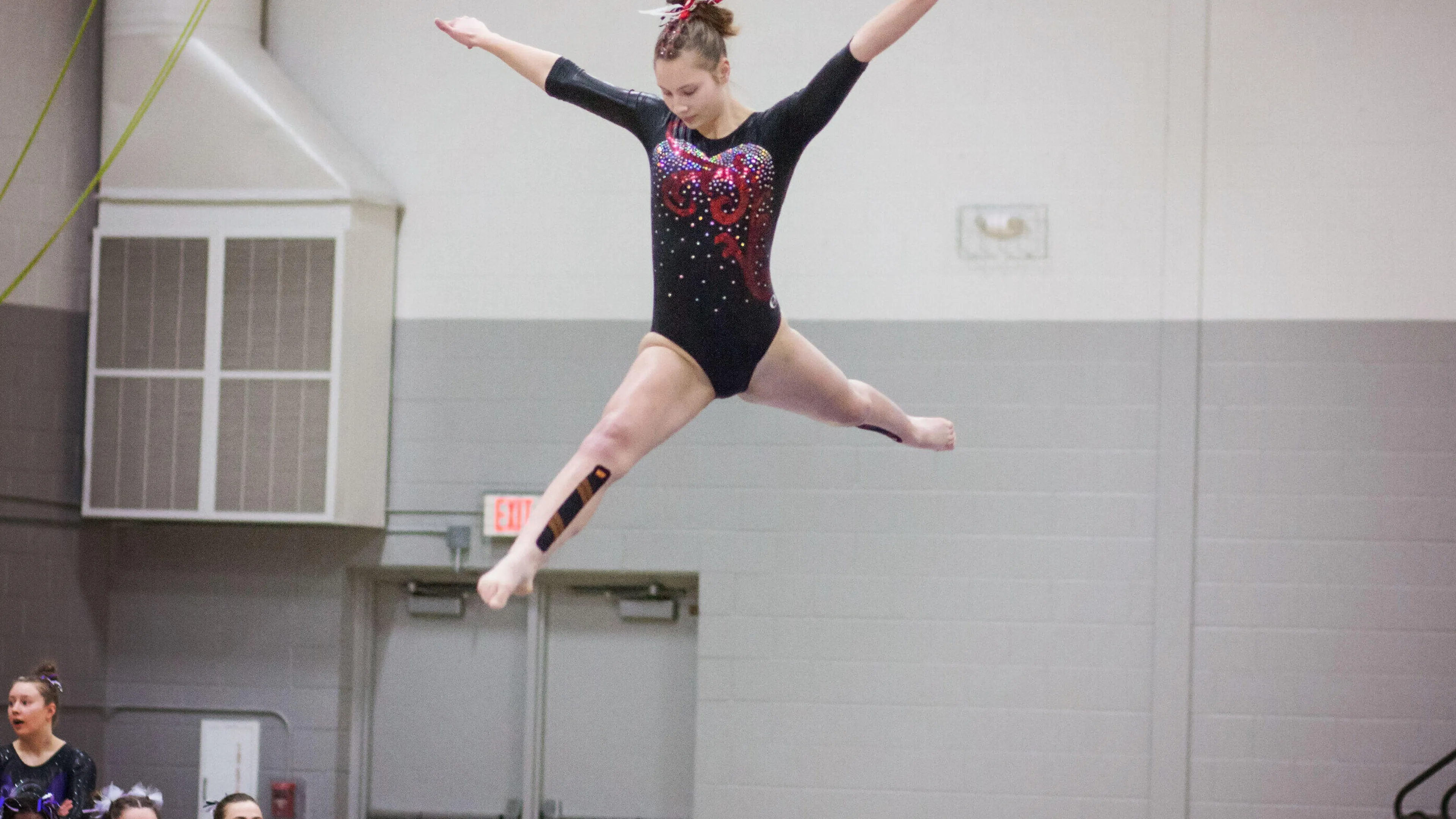 A young gymnast in a black leotard with red accents performs a graceful leap over a balance beam.  Spectators watch from the sidelines.