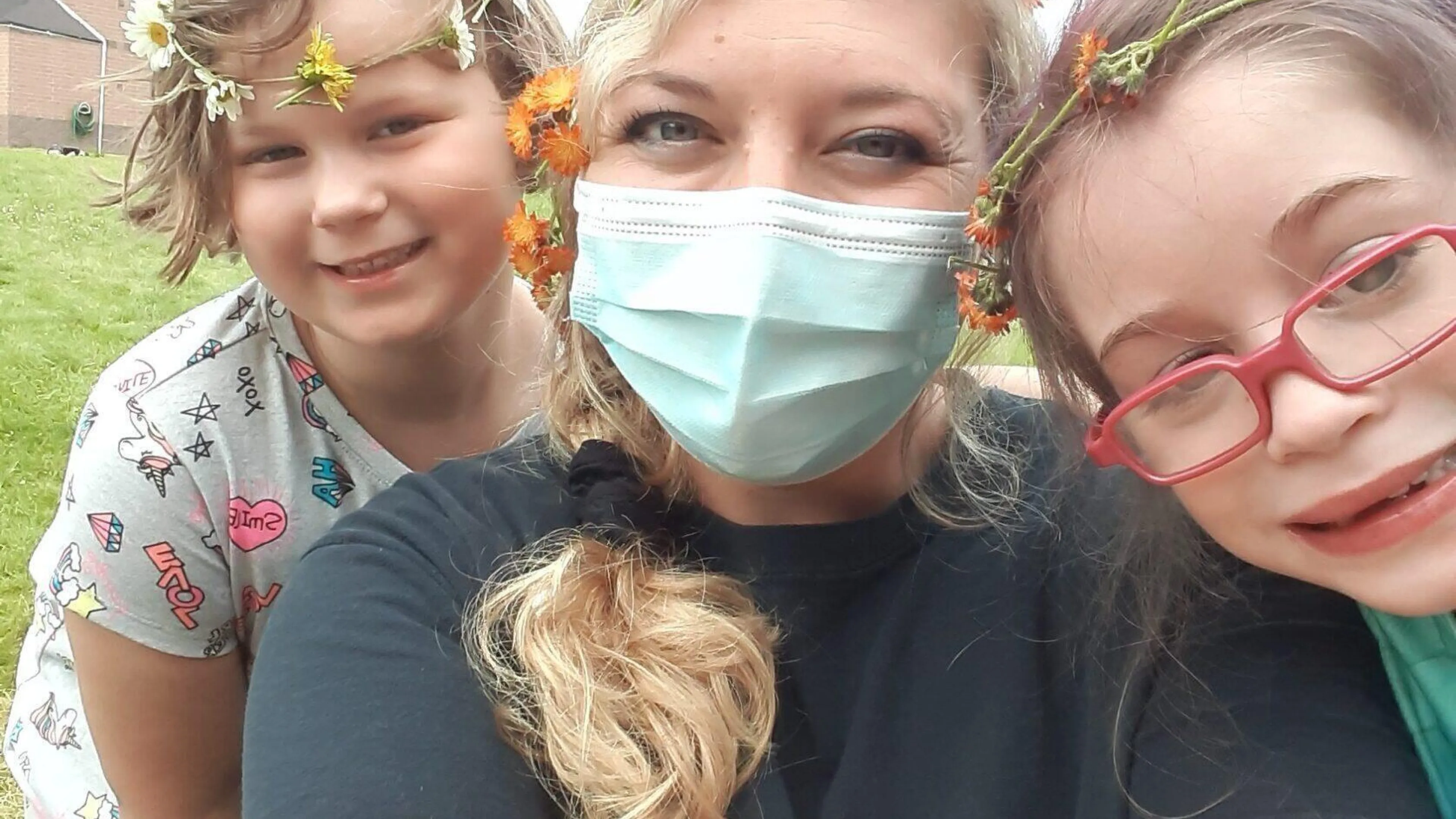 Smiling children and a counselor wear flower crowns outdoors.  The counselor wears a mask and a black t-shirt with mirrored text.  A fun summer camp moment.