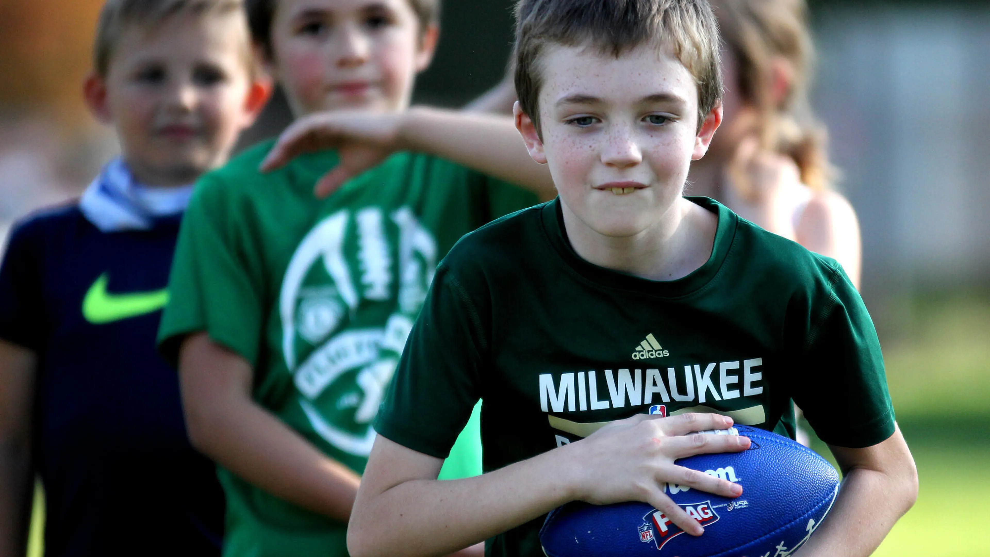 Kids in green shirts participate in a youth sports program; one boy holds a blue football.