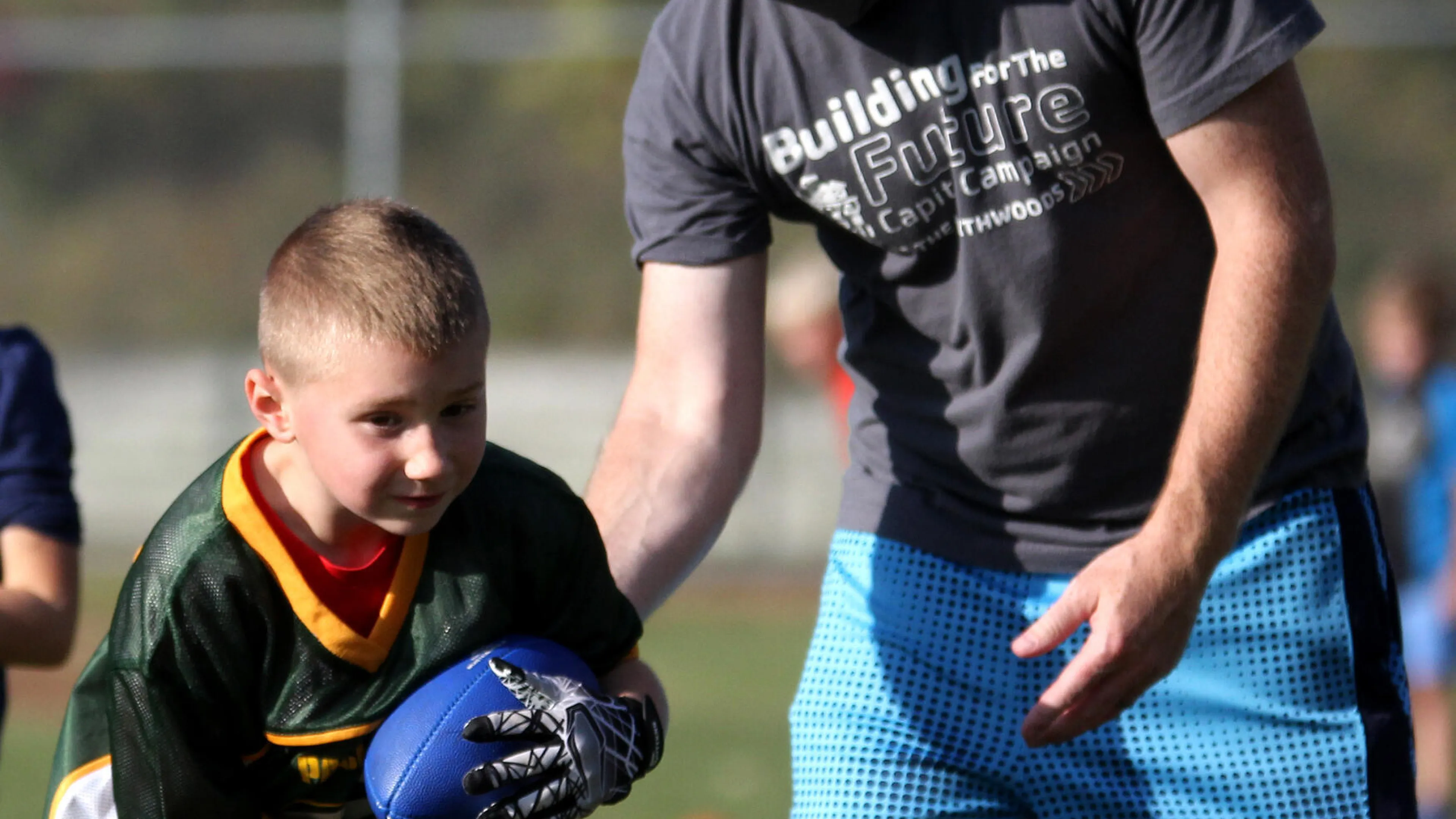 A young boy in a football uniform grips a ball, receiving coaching from a man wearing a face covering and a gray "Building for the Future" campaign shirt.  They're on a grassy field.
