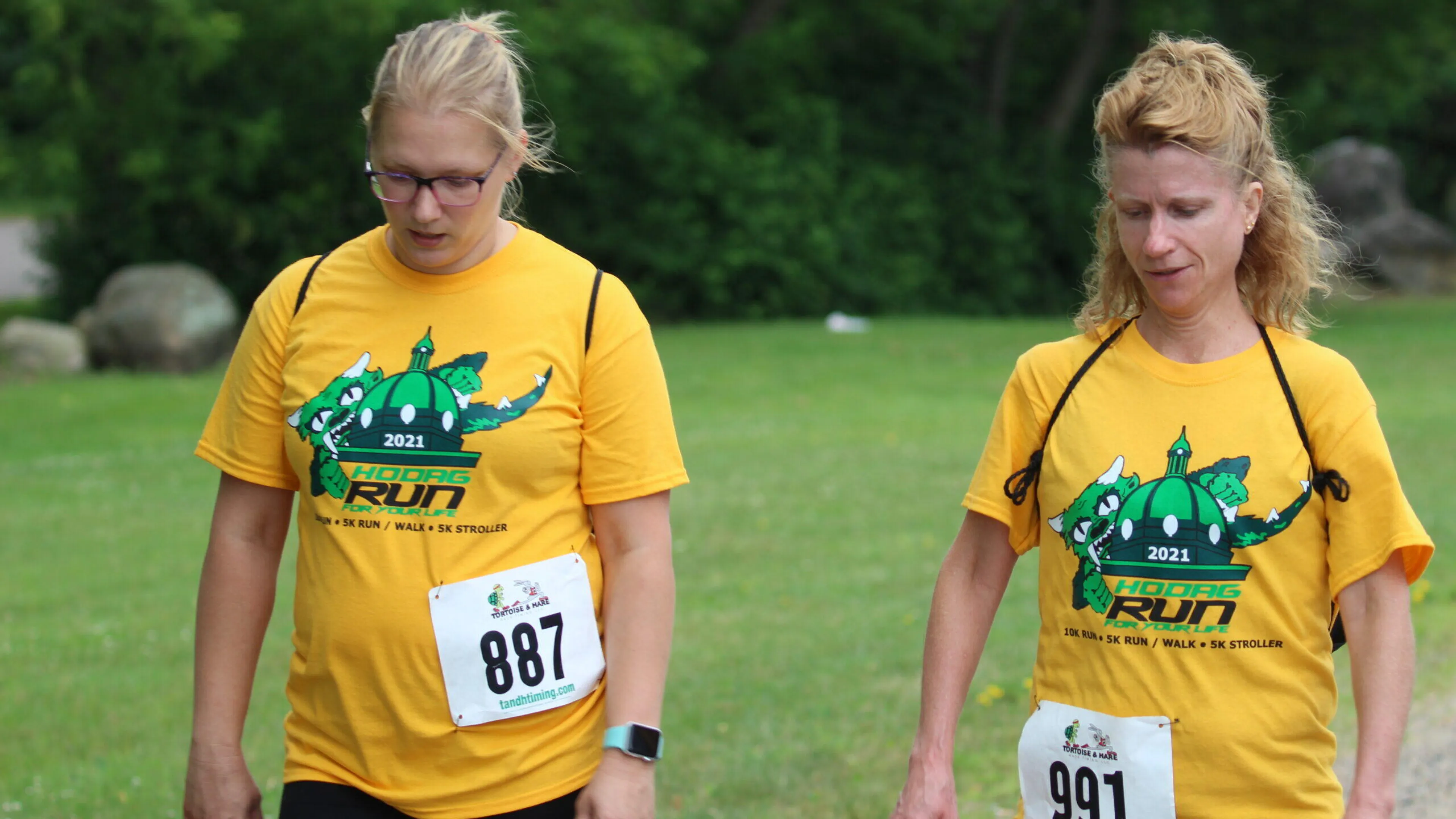 Two women in yellow t-shirts, bearing the 2021 Holcomb Run logo, walk outdoors.  Their race bib numbers are 887 and 991.