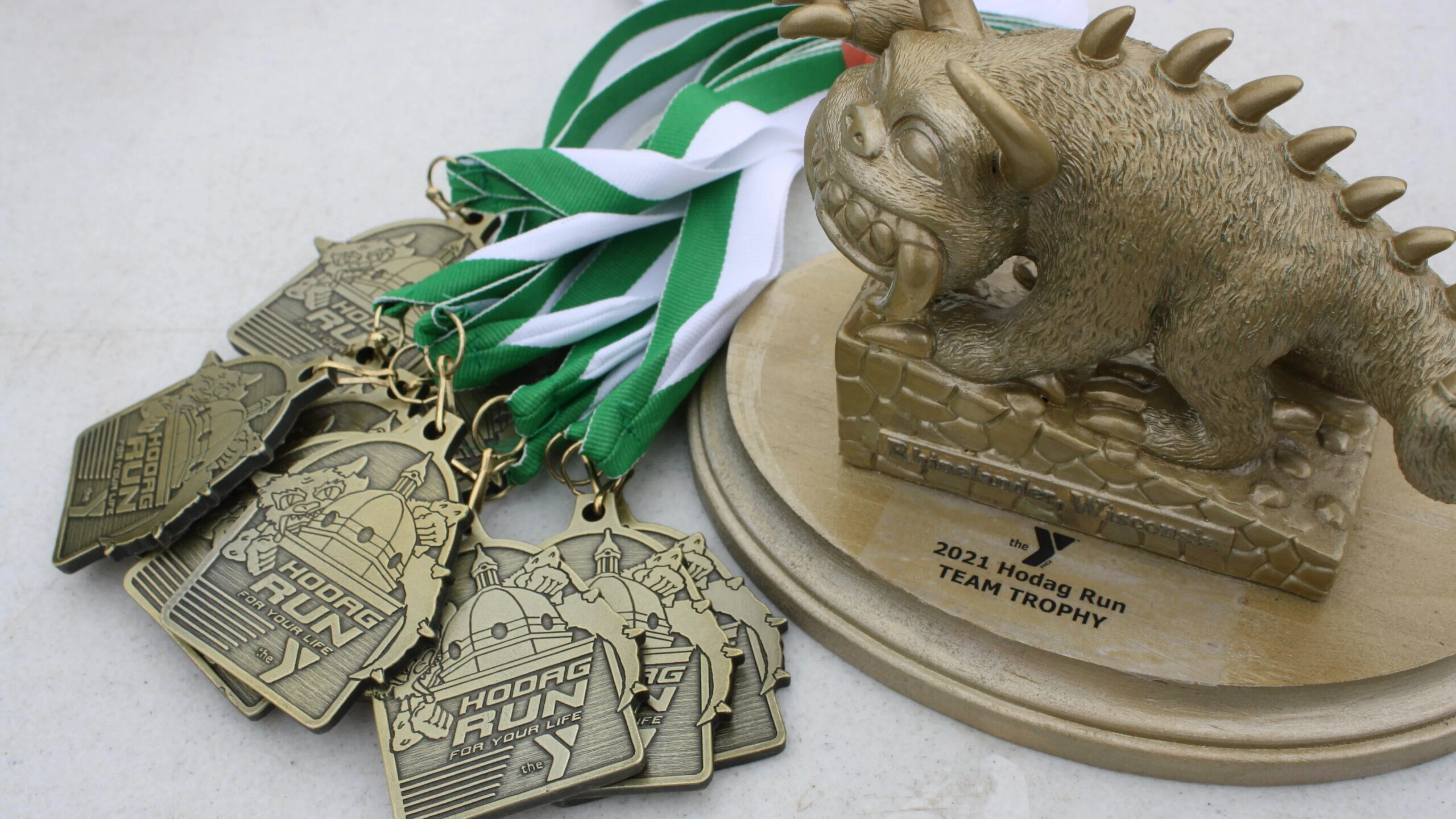 Several bronze medals from a Hodag Run,  alongside a gold team trophy featuring a sculpted Hodag.  Green and white ribbons are included.