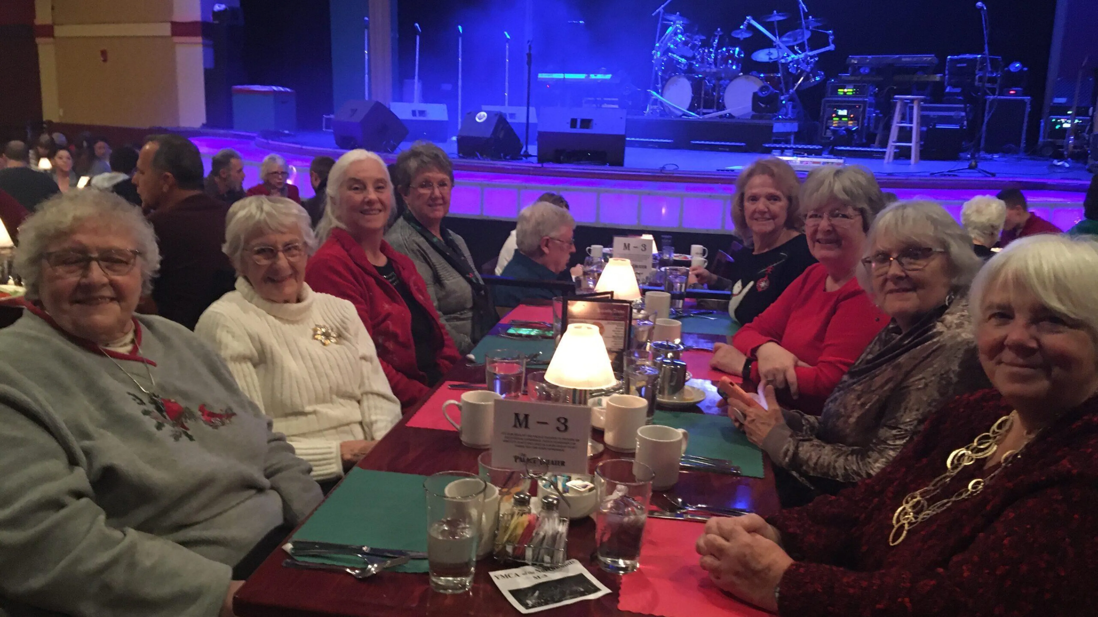 A group of women enjoy a meal together at a table in a theater.  A stage with musical instruments is visible in the background.