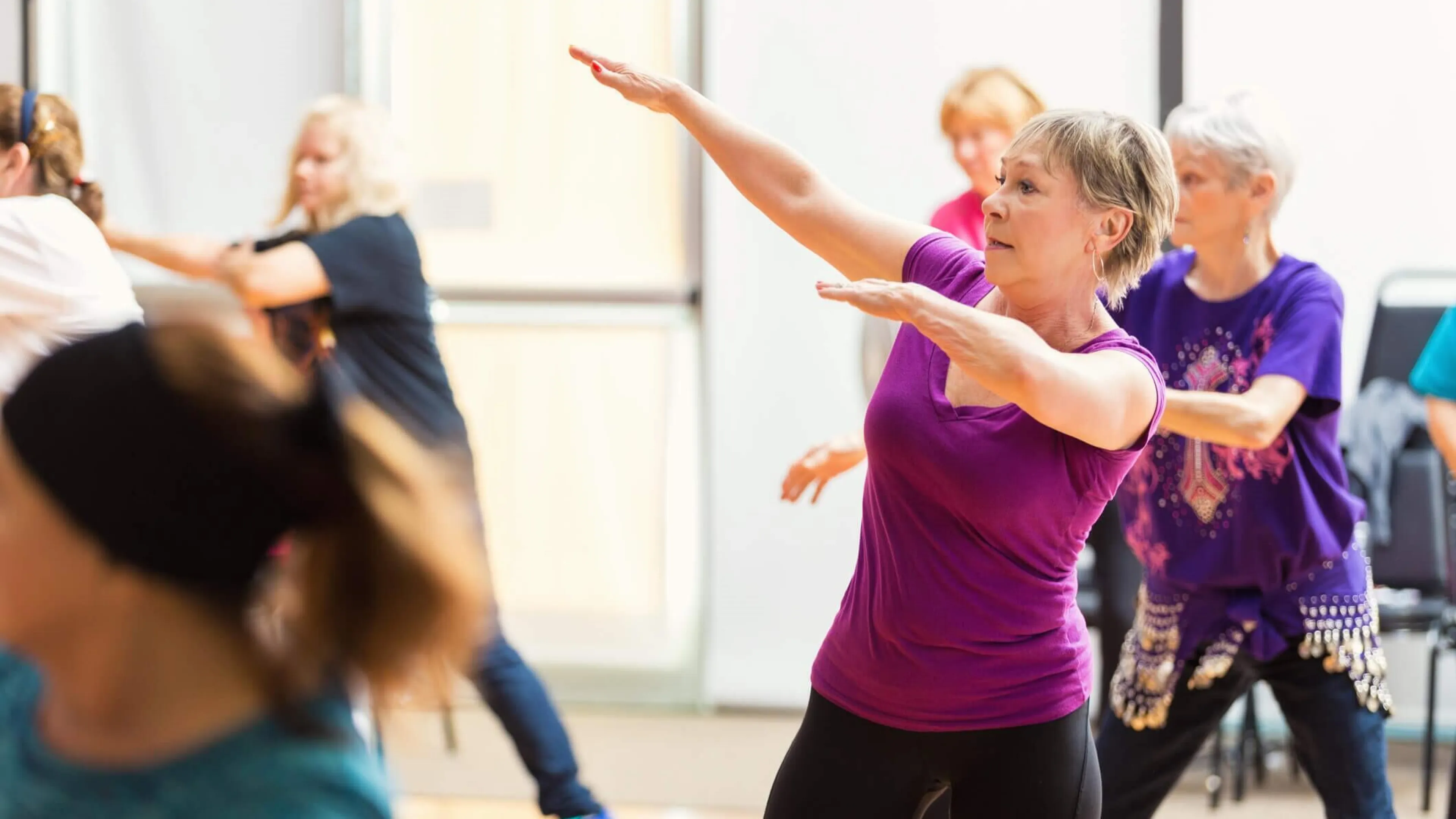 Women of varying ages participate in a fitness class, gracefully moving their arms and bodies in unison.  One woman in a purple shirt is prominently featured, her arms outstretched.