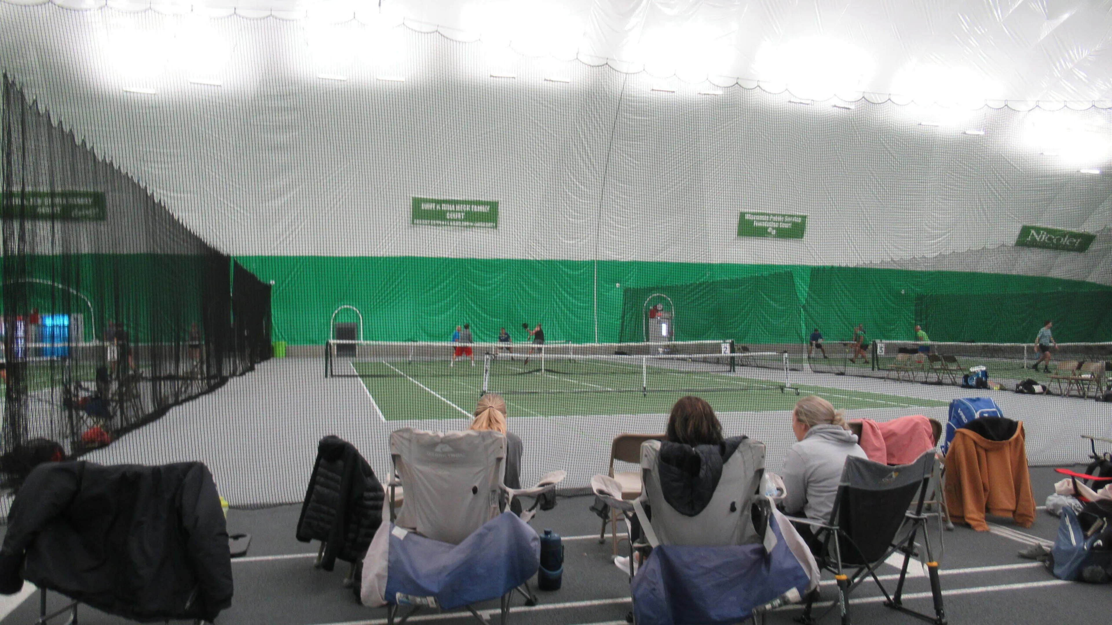 Indoor tennis court with spectators seated in lawn chairs watching a tennis match.  Several children are playing on the court.