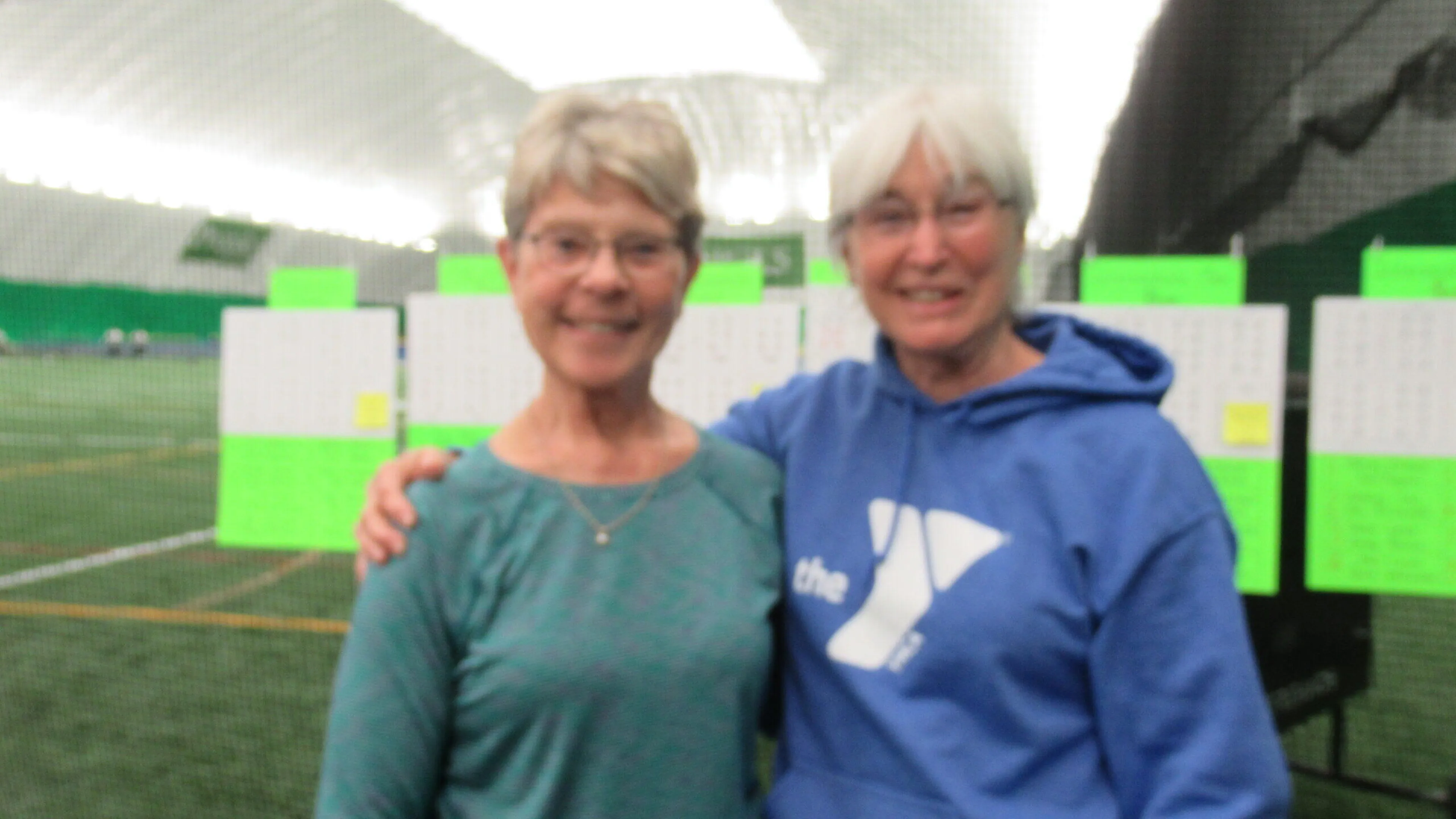 Two women stand together in an indoor sports facility; one wears a teal shirt, the other a blue hoodie.  Green scoreboards are visible in the background.