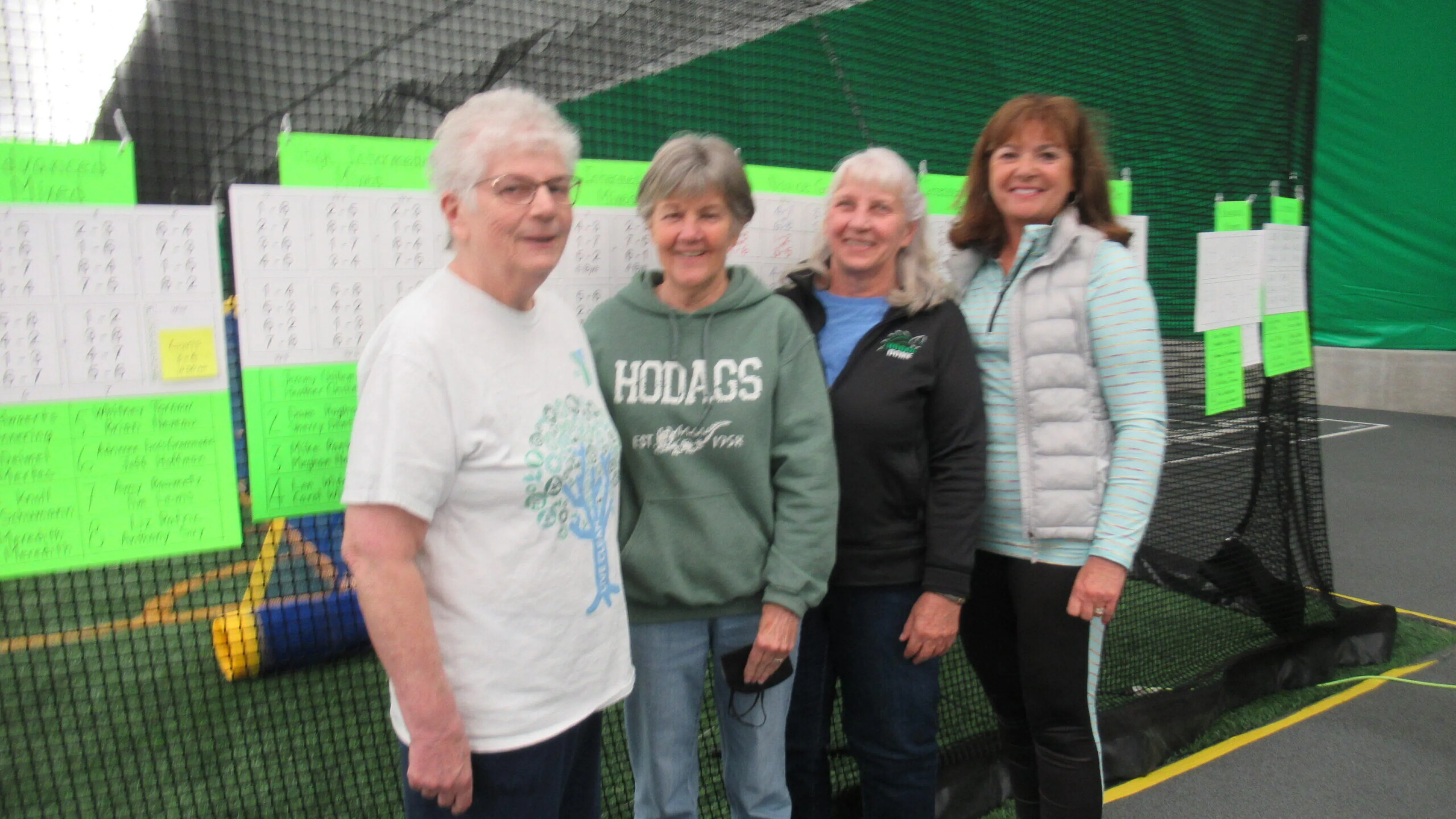 Four women stand smiling near score sheets; one wears a green sweatshirt, another a white shirt, and two others have on darker jackets.  They're in an indoor sports facility.