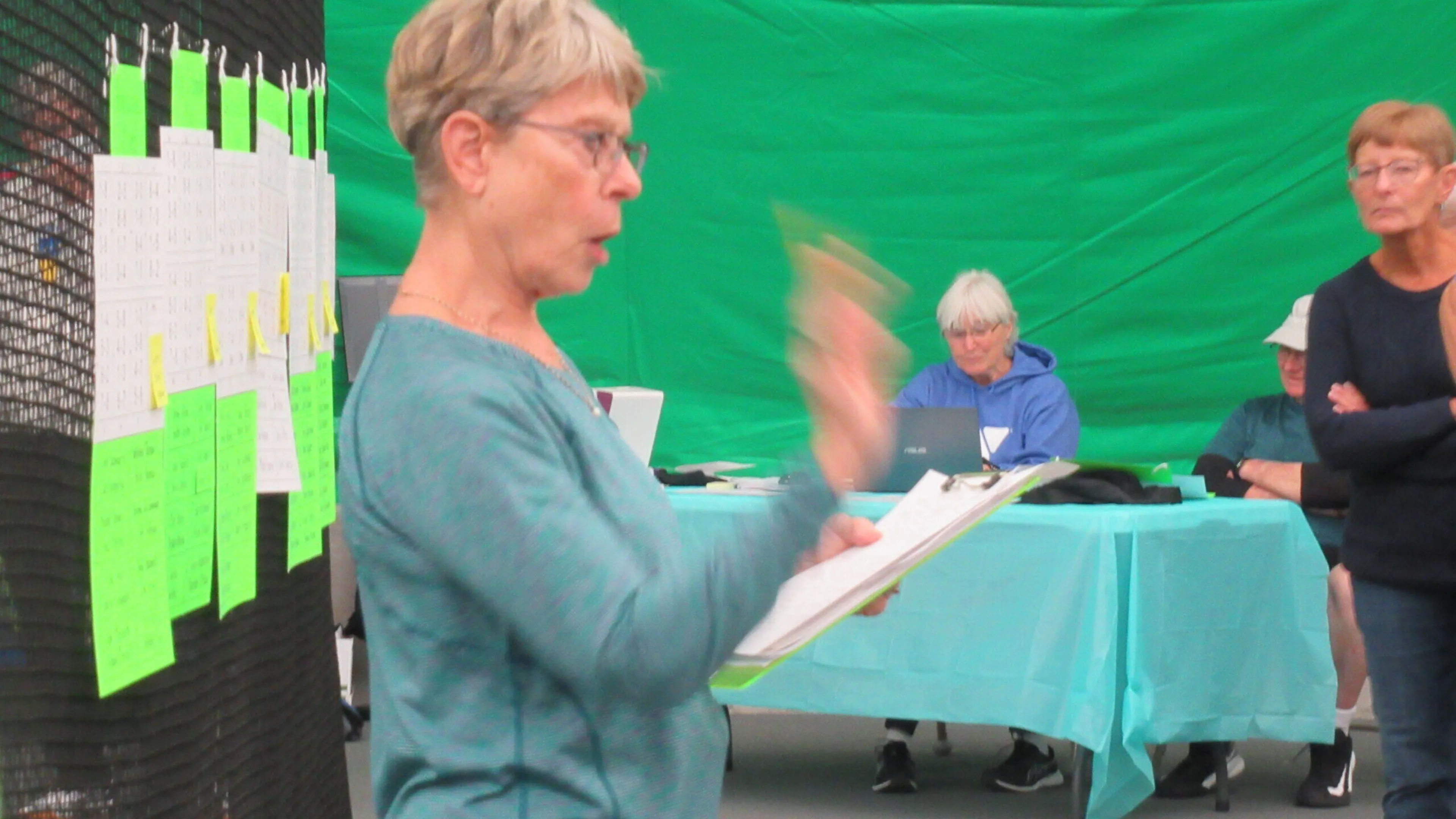 A woman in teal, holding a clipboard, addresses a group.  Bright green backdrop.  Others are seated at a light blue table, some with laptops.  Notes hang on a black mesh screen.