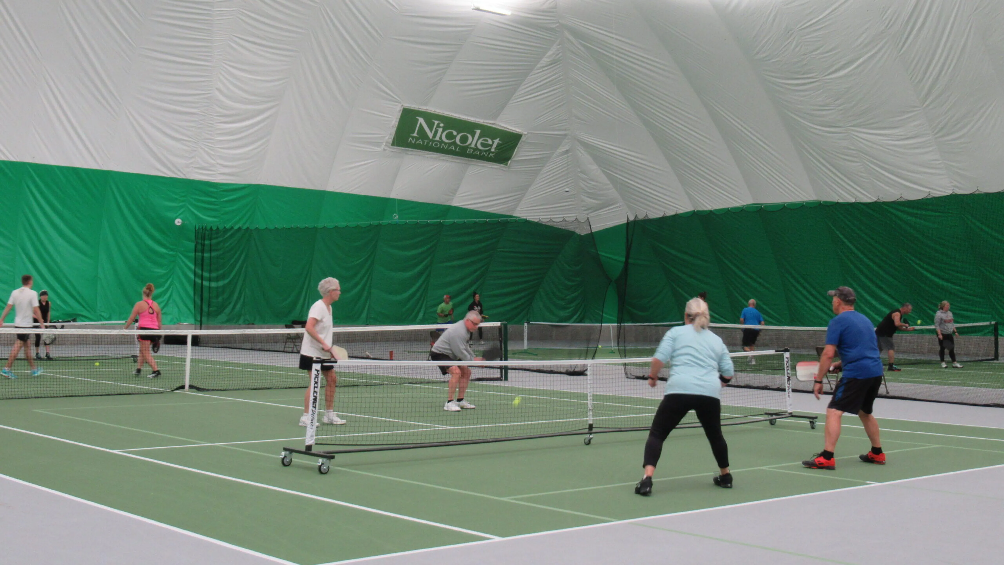 Active adults playing pickleball indoors on several courts.  Nicolet Athletic Center banner visible.