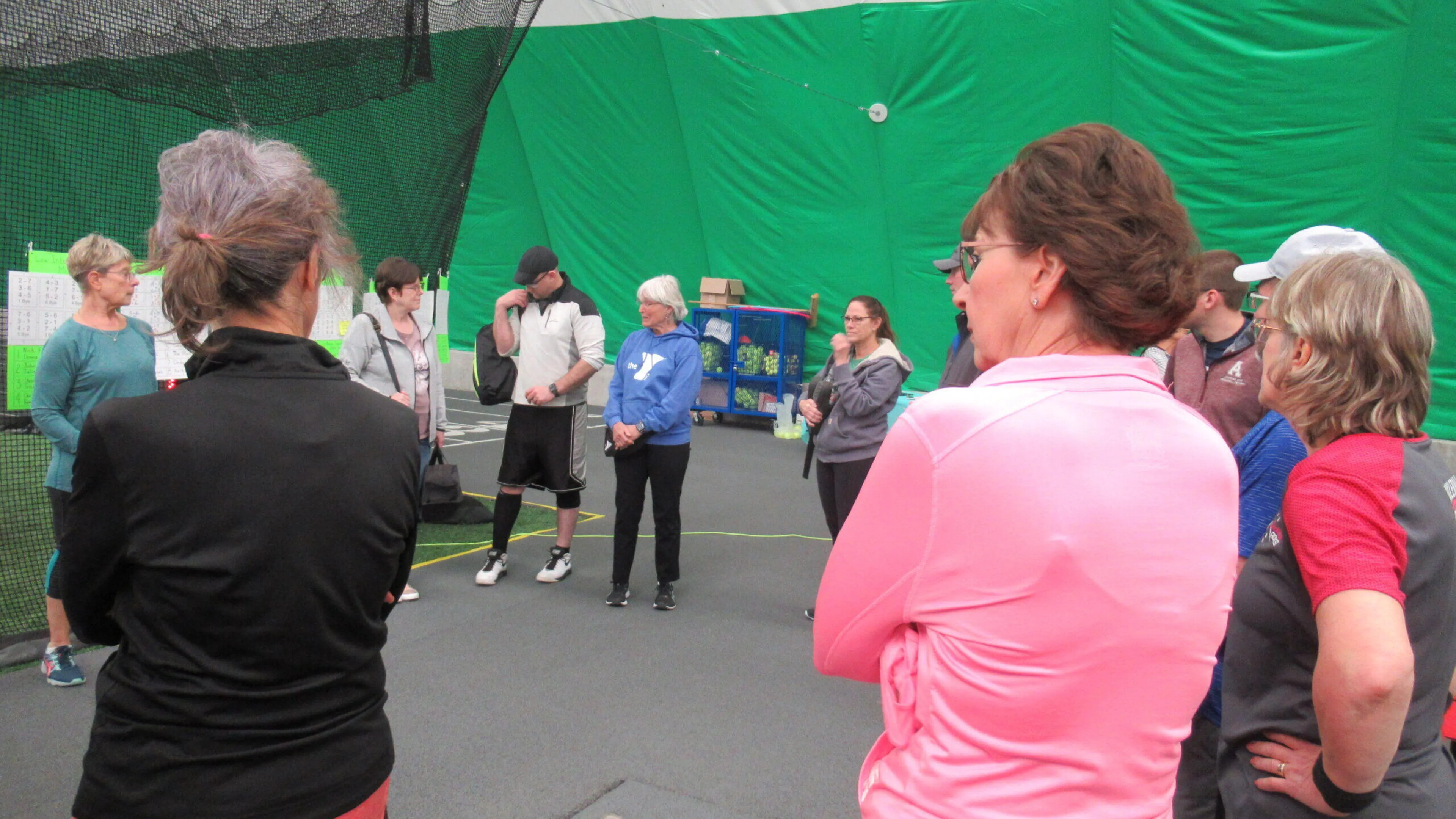 A group of adults stand in an indoor facility, some appearing to listen to instructions.  A green wall is in the background.