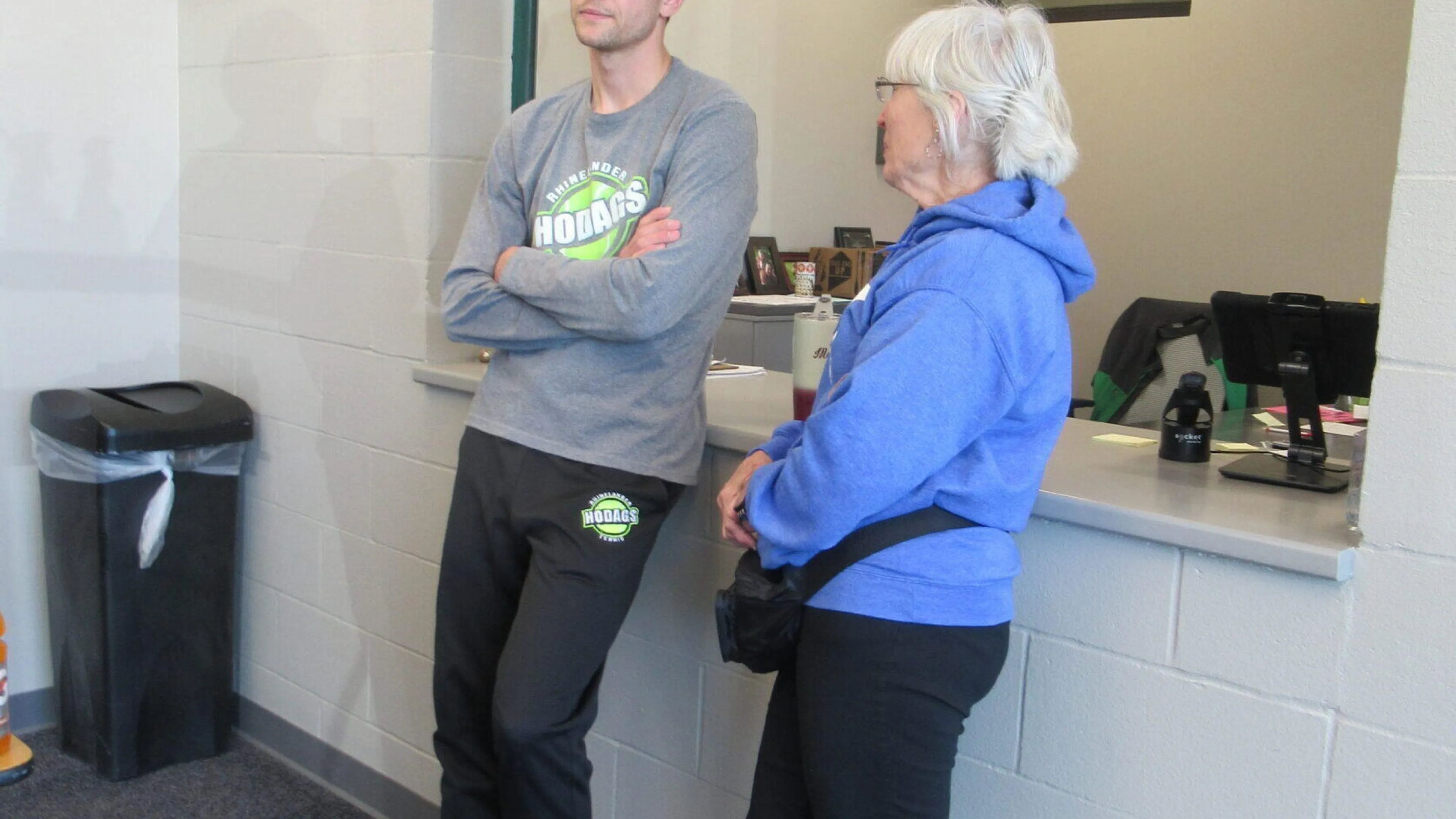 A young man and older woman stand at a counter, talking.  He wears a gray sweatshirt and black sweatpants; she's in a blue jacket and black pants.