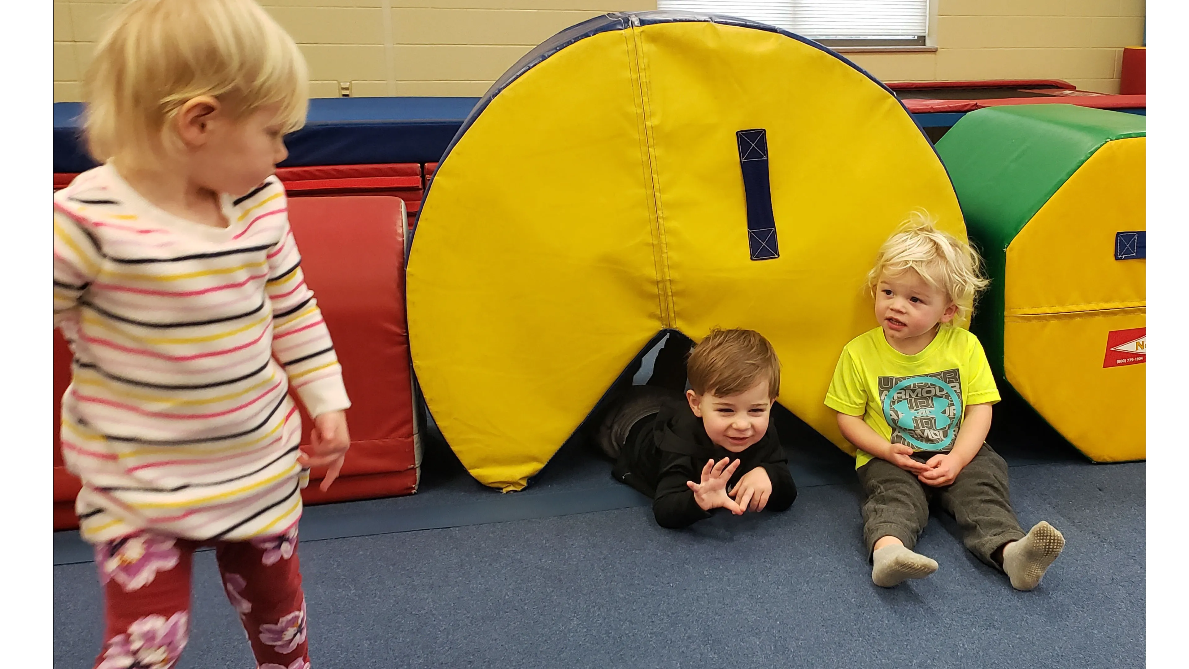 Three toddlers playing in a gym; two boys are near a yellow play tunnel, and a girl stands nearby.