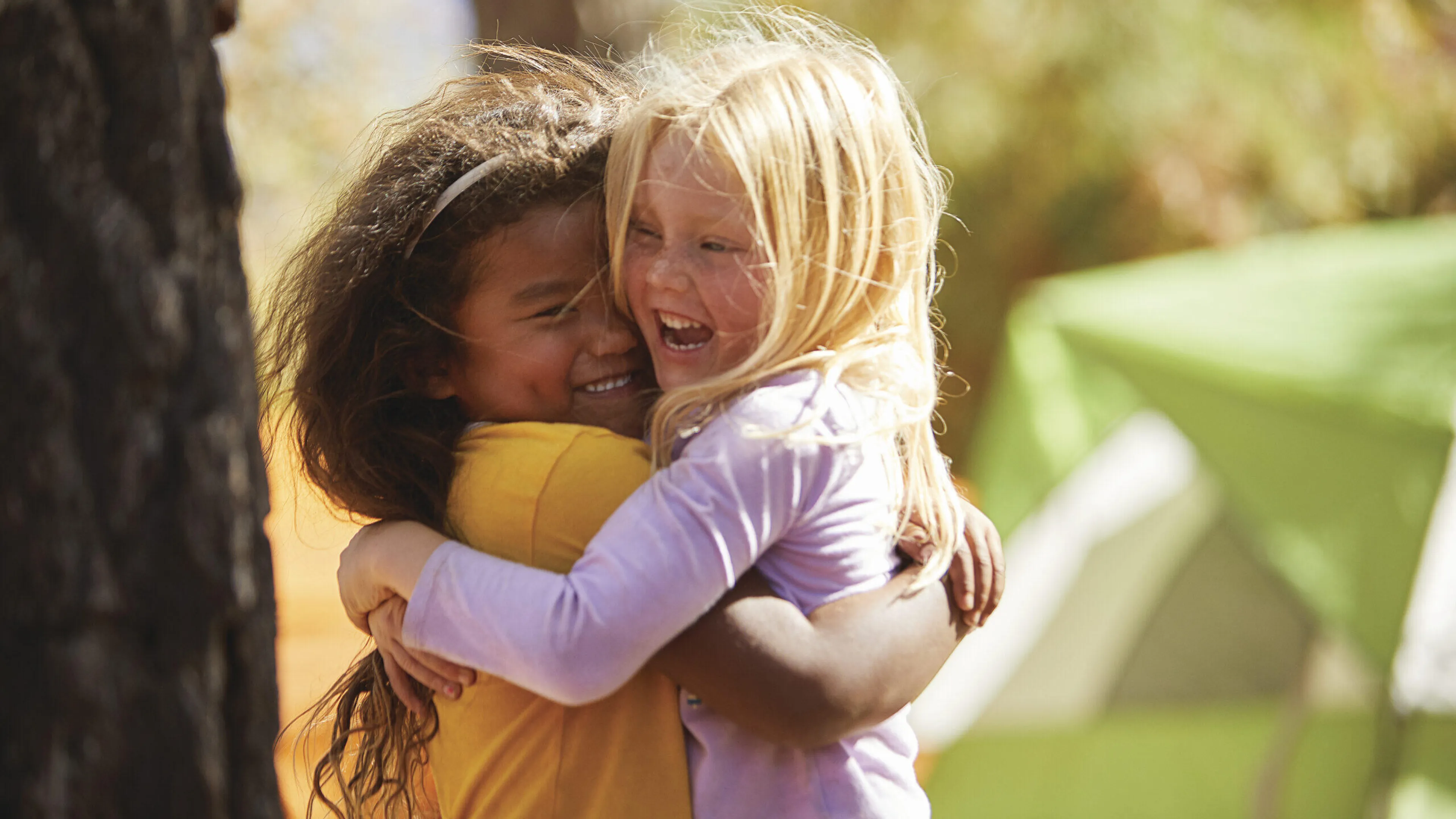 Two girls hug joyfully at a summer camp,  one in a yellow shirt and the other in lavender.
