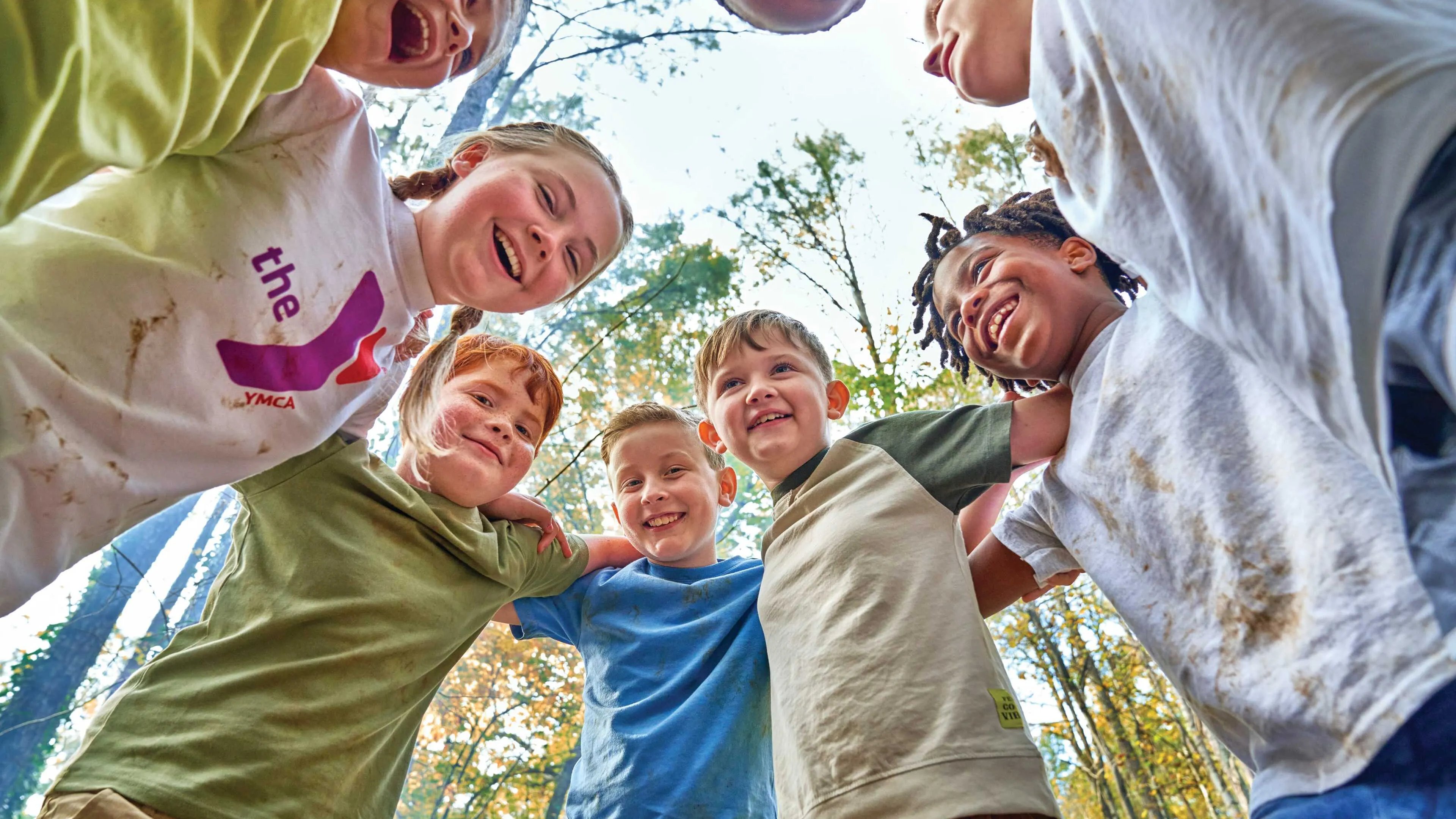 Happy children, arms around each other, smiling at the camera in a wooded area.  They appear to be participating in an outdoor program.