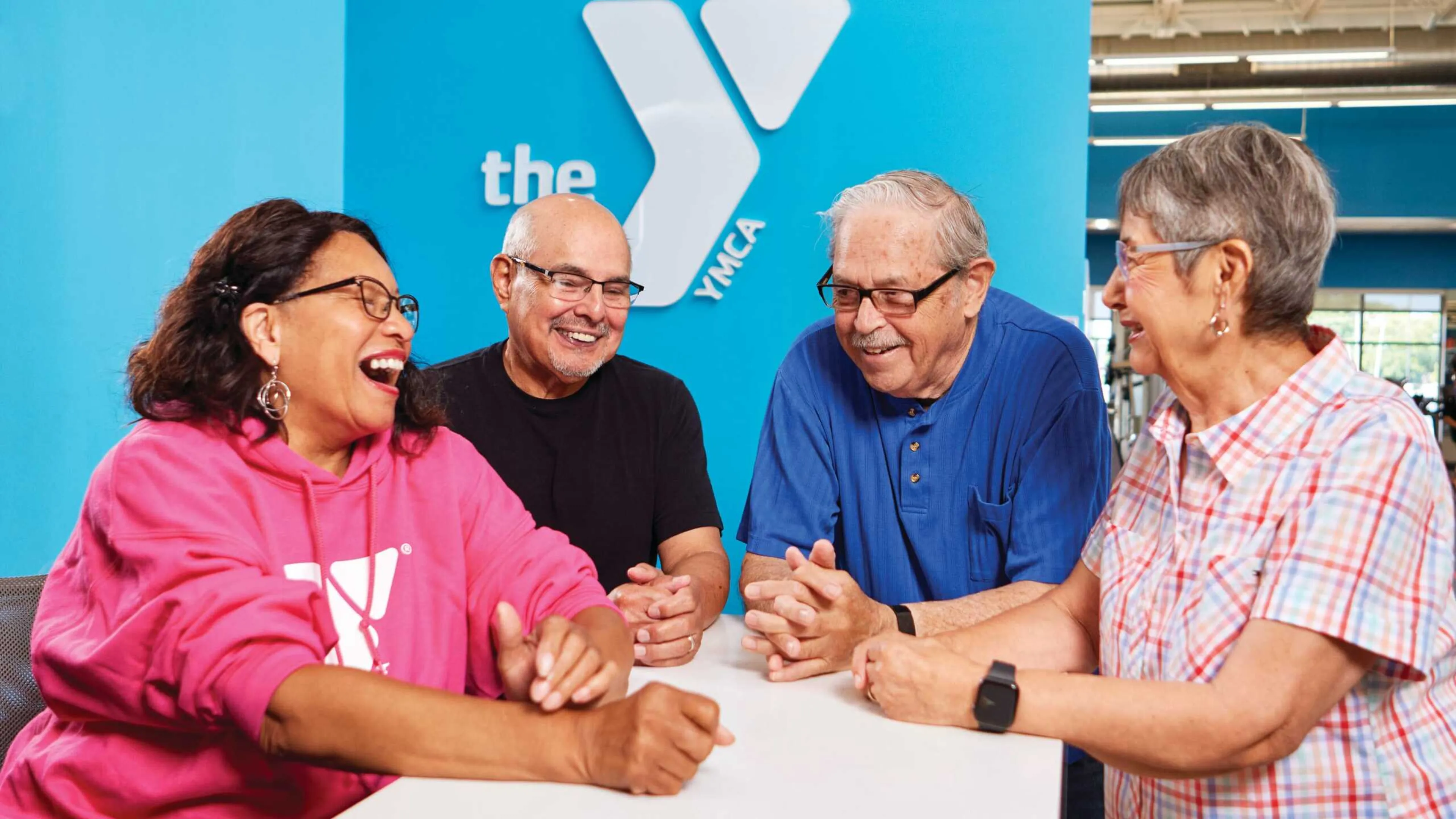 Four adults laugh together at a long white table against a bright blue wall with a logo.