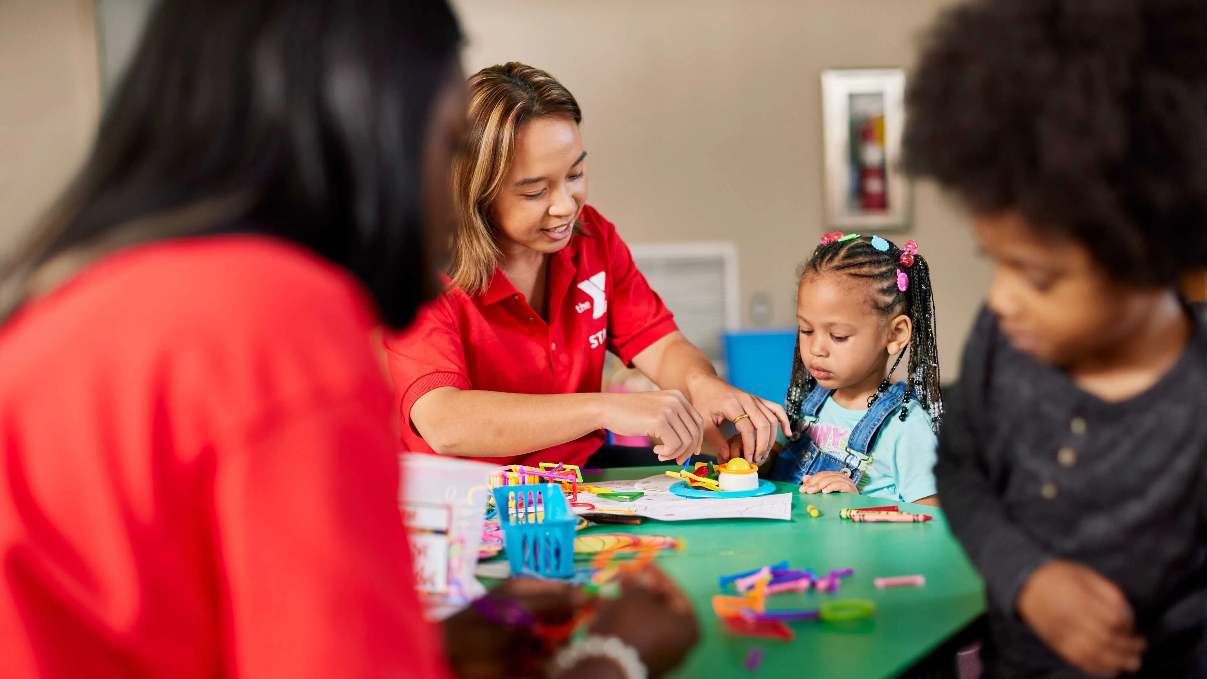 A female counselor assists a young girl with a craft project at a kids' activity table.  Another child sits nearby, and a second adult is visible in the background. Colorful art supplies are scattered on the table.