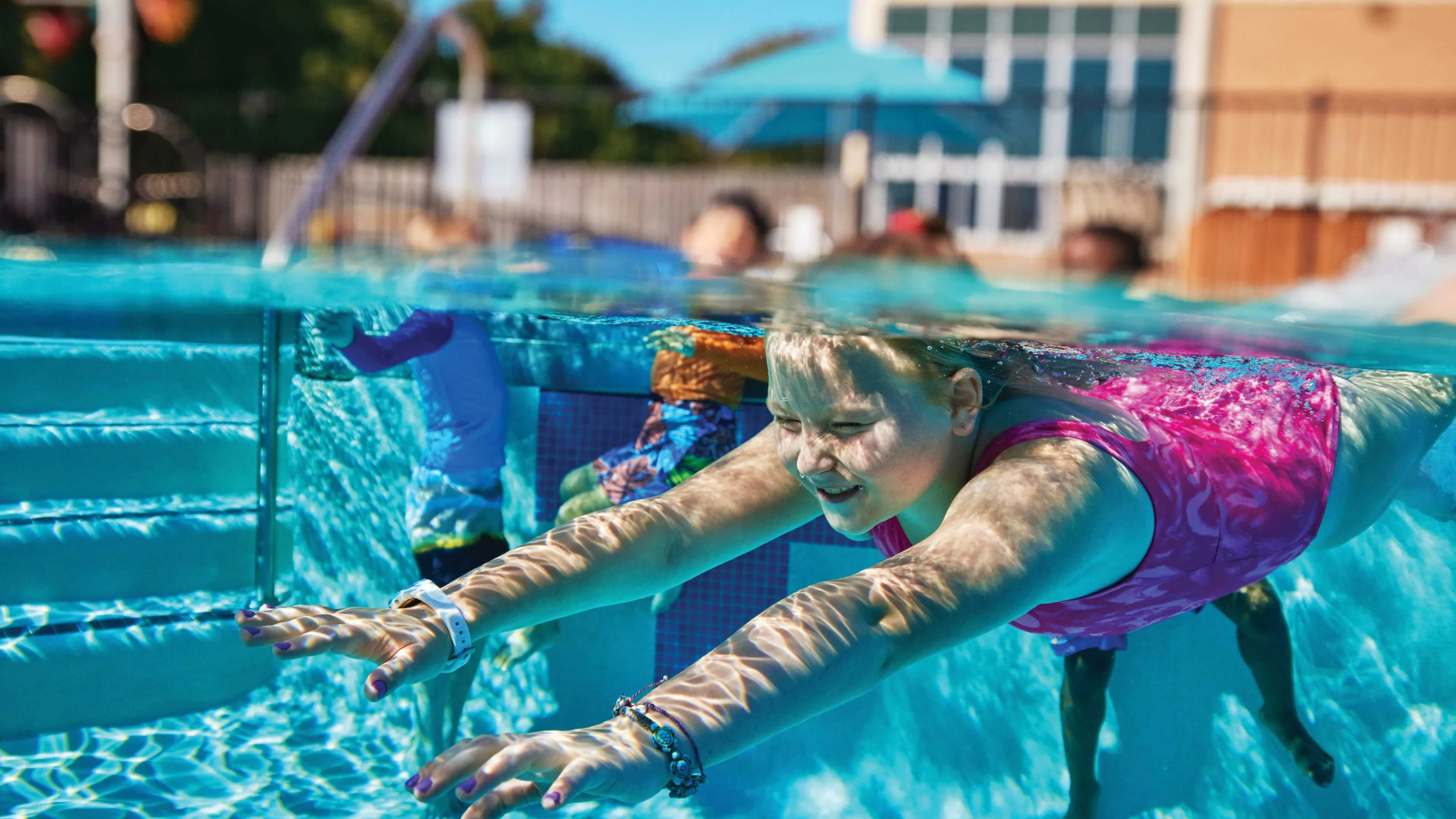 A girl in a pink swimsuit swims underwater in a pool; other children are visible in the background.