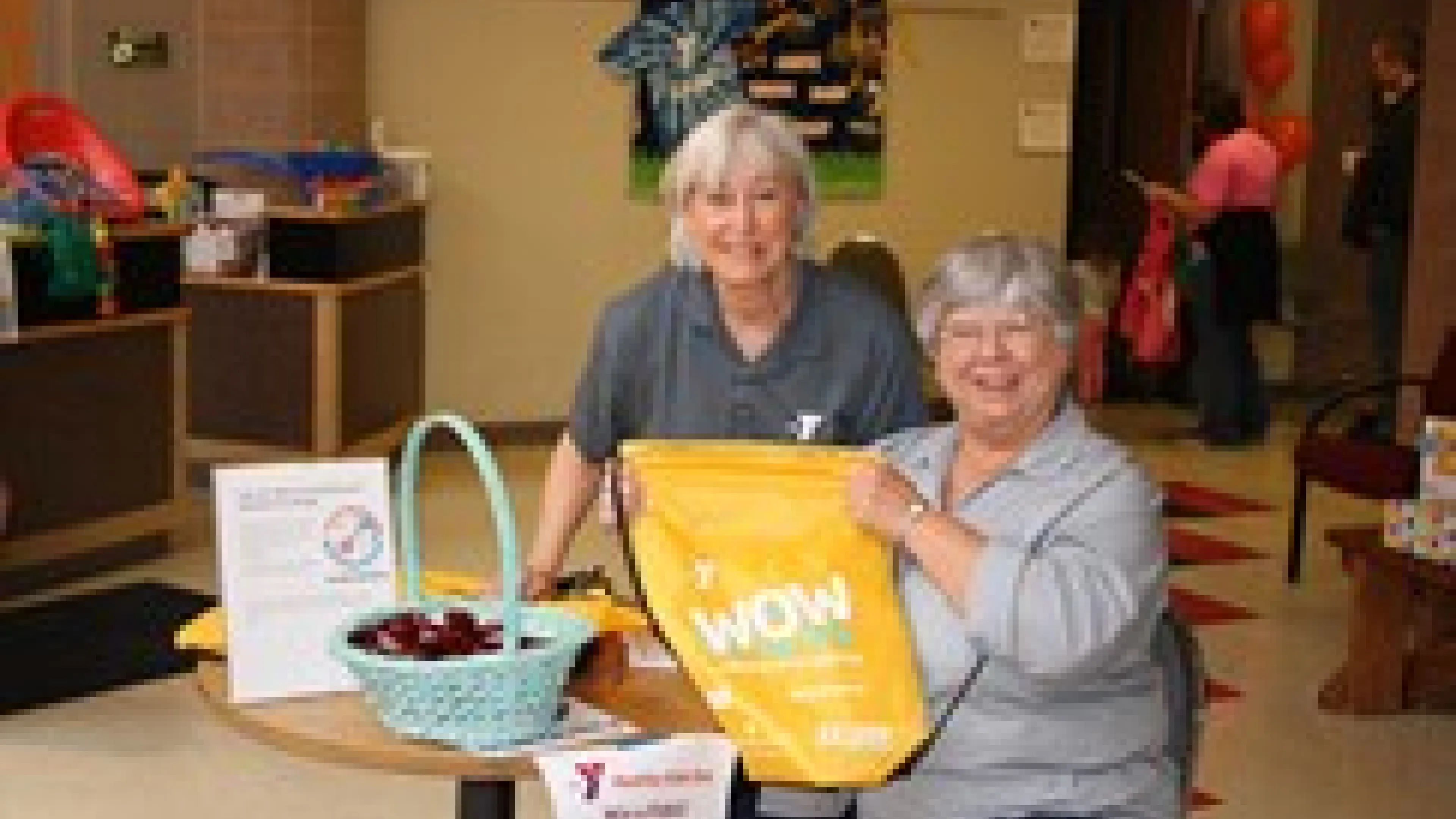 Two smiling women stand behind a table filled with treats and information at a community center event. The lady holds a yellow tote bag labeled "WOW".