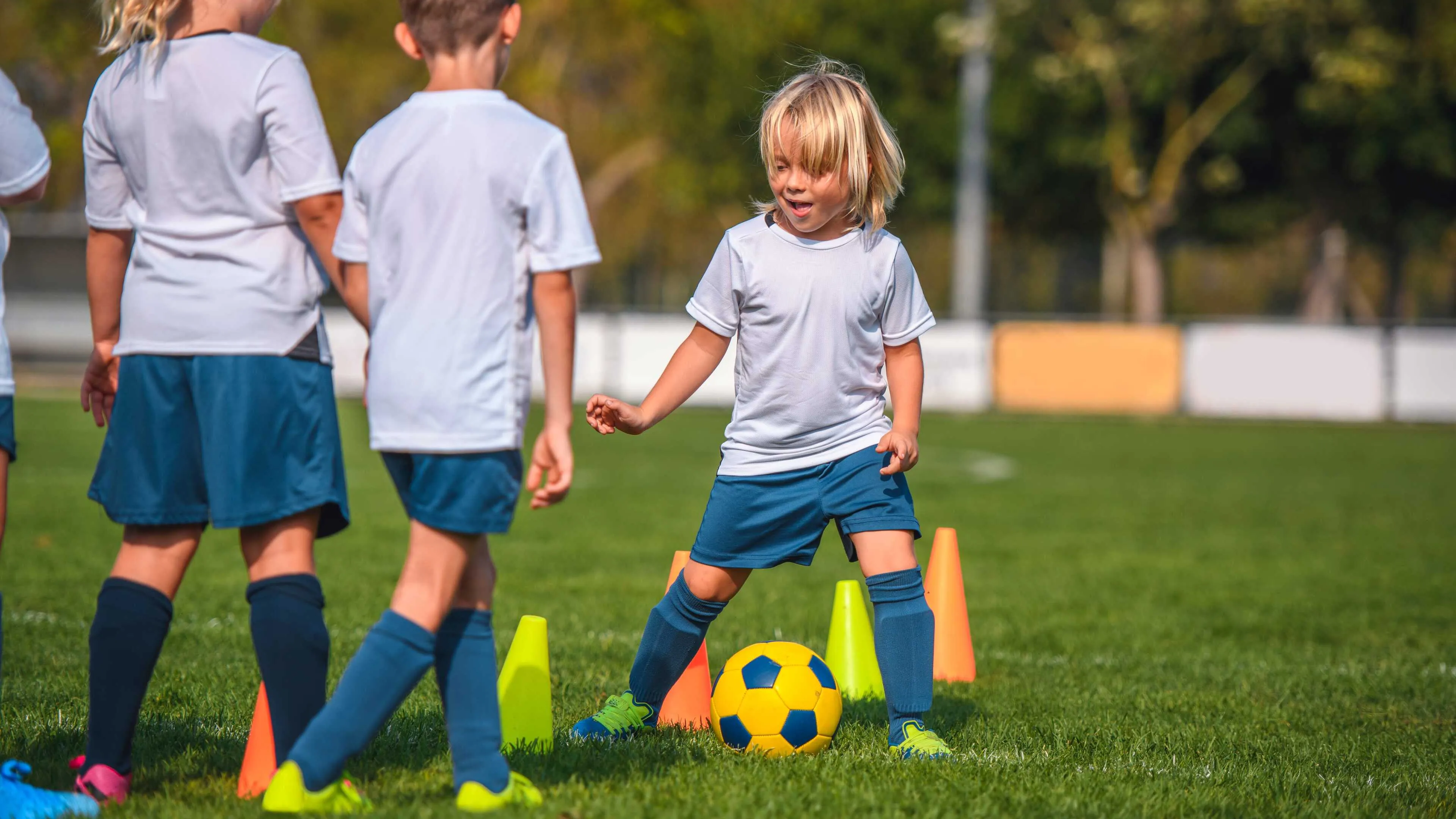 Kids in white soccer jerseys and navy shorts practice dribbling a soccer ball around cones on a grassy field.