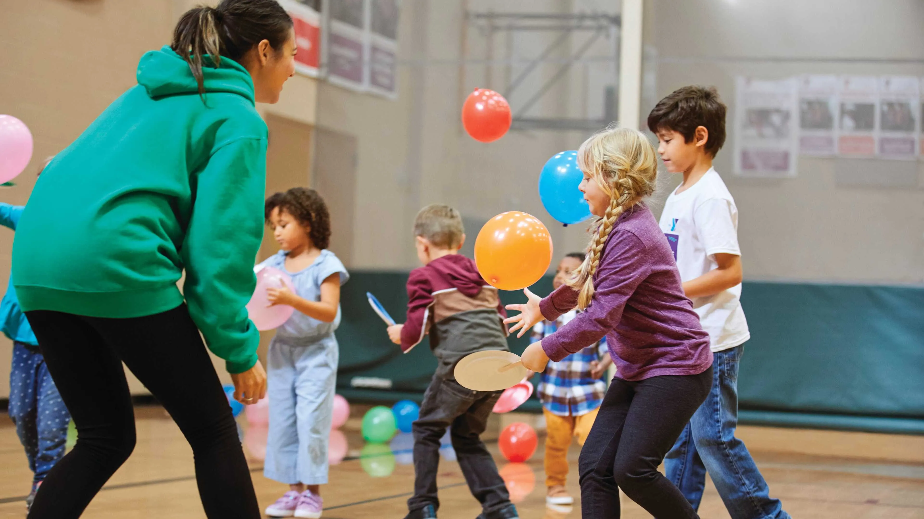 Kids play balloon games in a gym with a counselor.  Colorful balloons float, children use paddles, and smiles abound.
