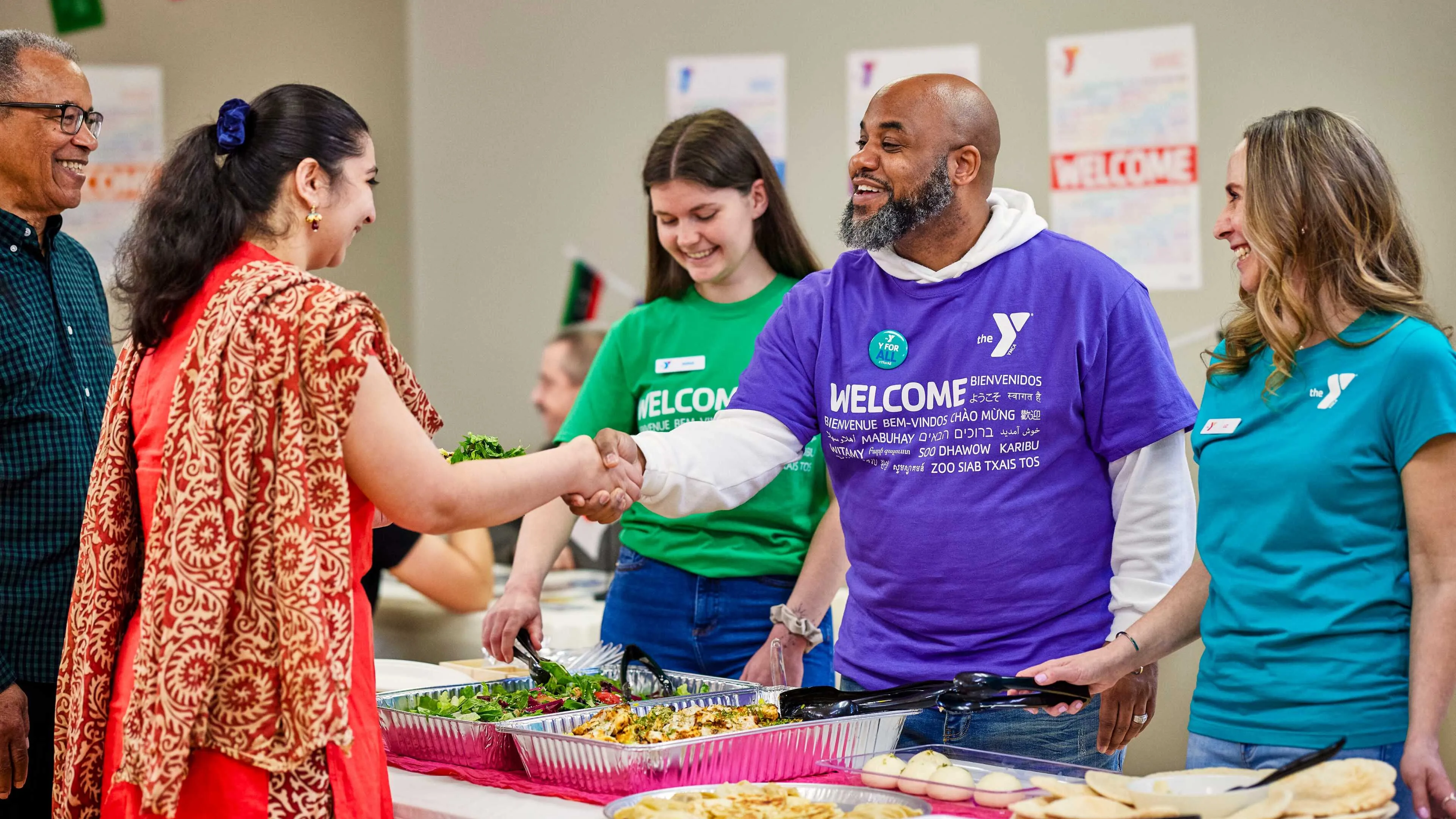 Smiling people shake hands at a community event; food is on tables decorated with international flags.