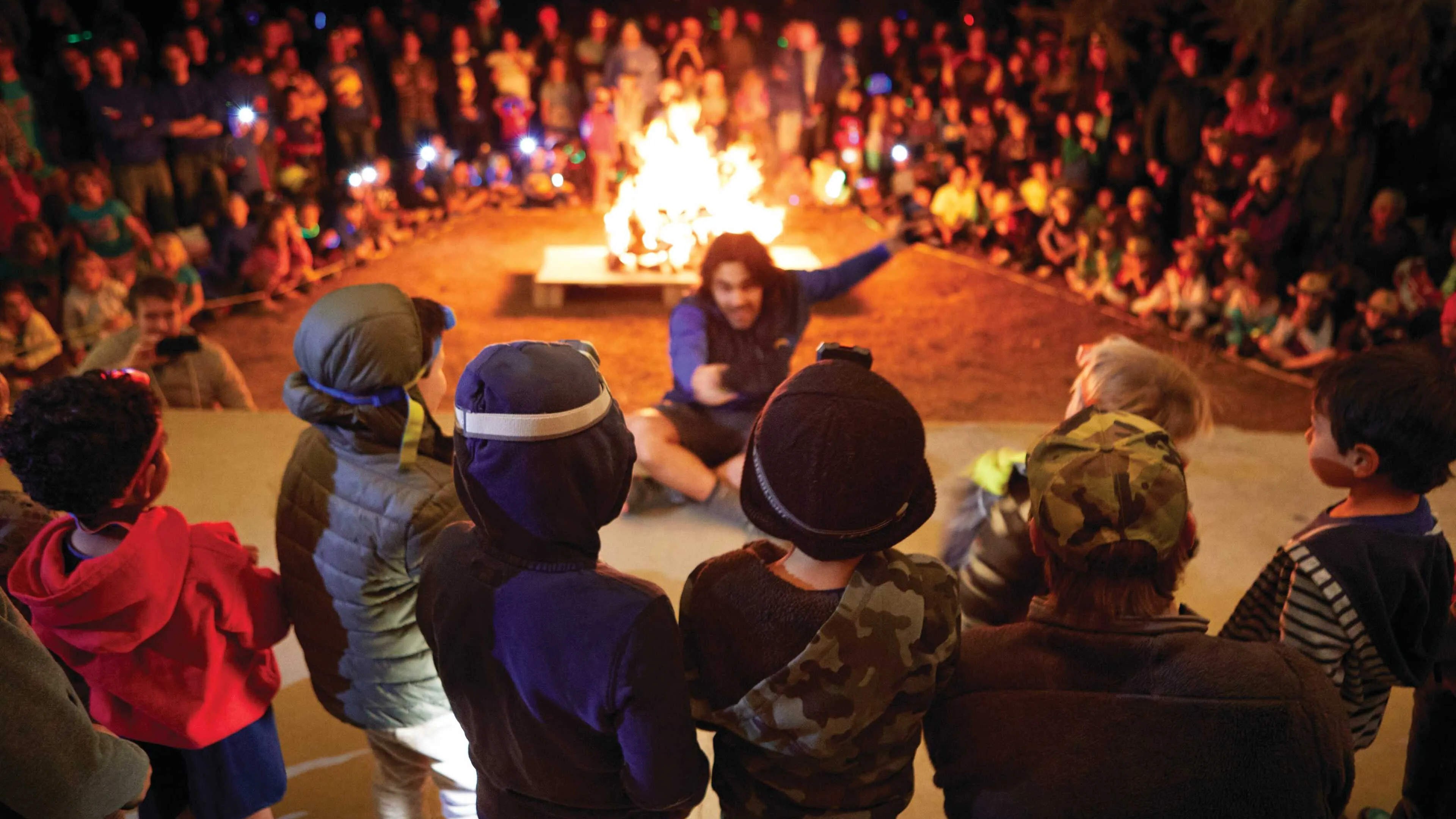 Children gather around a bonfire, captivated by a storyteller.  A large crowd watches from behind the kids. Warm light illuminates the scene.