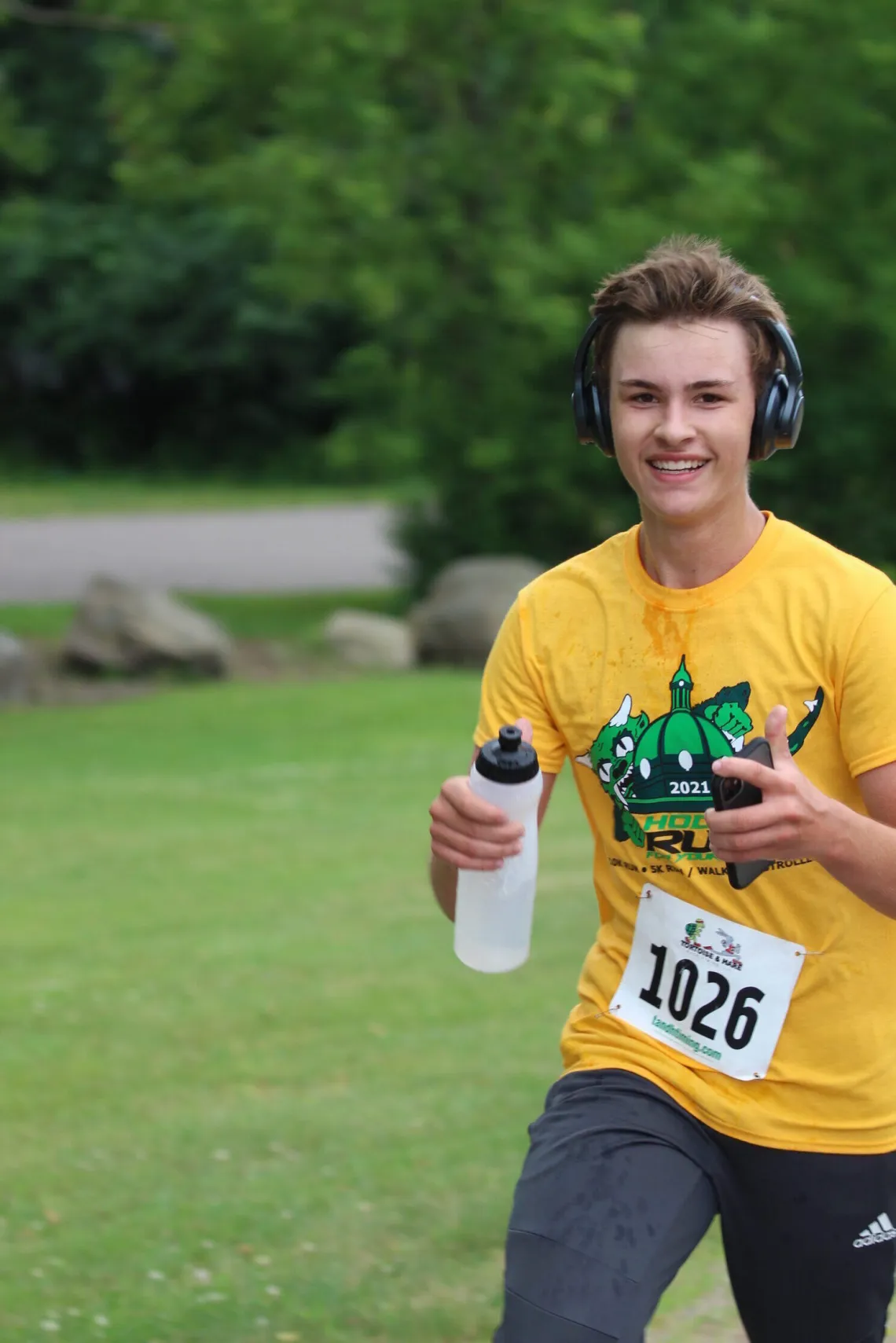 Smiling teen in a yellow race t-shirt and headphones, holding a water bottle, runs a race, bib number 1026.
