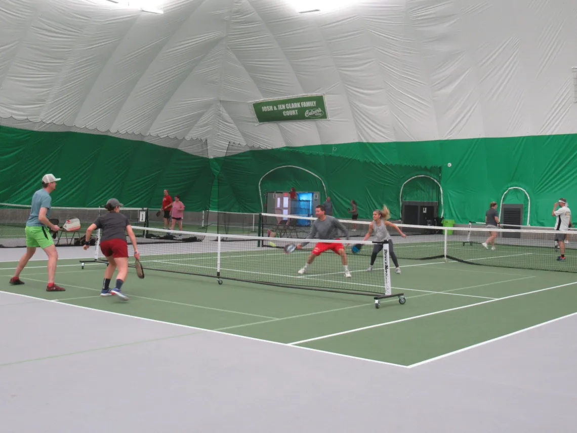 Several people play pickleball on an indoor court.  Some wear hats; others are wearing athletic clothing.  The court is green with white lines.
