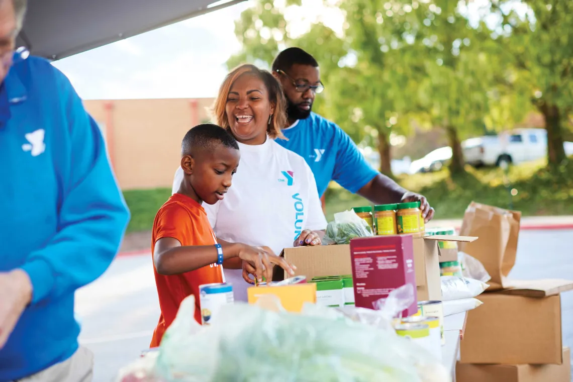 Smiling volunteers, including a boy, sort food donations into boxes.

