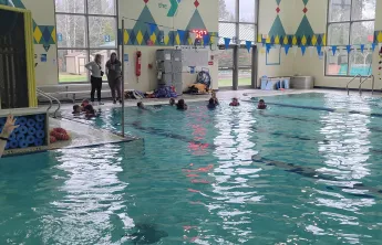 Kids participate in a swim lesson; instructors watch from the pool's edge.

