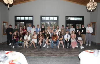 A large group of teens and young adults, many holding awards, pose for a group photo in a bright room.  They are seated in chairs arranged in rows.
