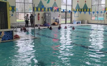 Kids participate in a swim lesson; instructors watch from the pool's edge.
