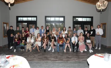 A large group of teens and young adults, many holding awards, pose for a group photo in a bright room.  They are seated in chairs arranged in rows.
